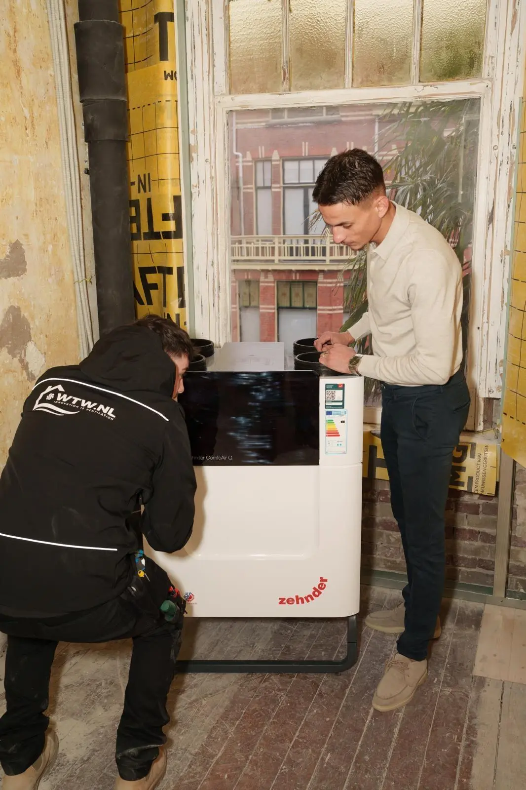 Moncif Hakmaoui, founder WTW.nl, squatting next to a Zehnder ComfoAir Q HRV unit